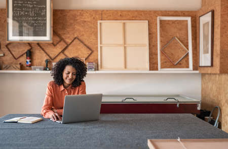Smiling young woman with curly hair using a laptop while working at a table in her picture framing shopの写真素材