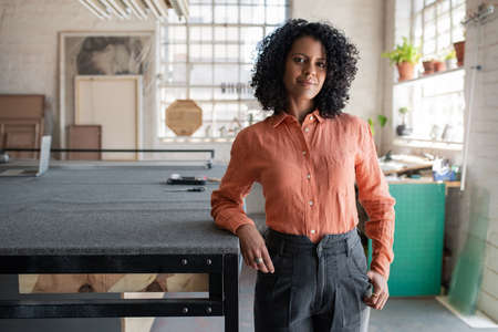 Portrait of a smiling young woman leaning against a large table in the work area of her picture framing studioの写真素材