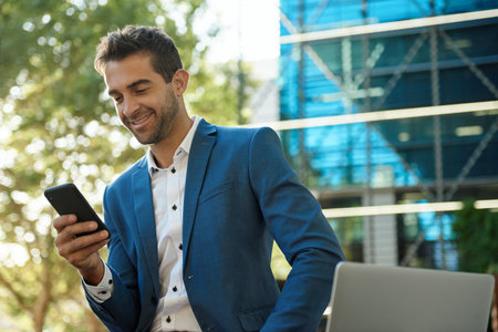 Smiling young businessman reading a text message on his cellphone while sitting outside of his office buildingの写真素材