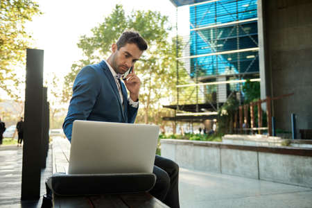 Businessman using a cellphone and laptop outside his office buildingの写真素材
