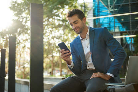 Smiling businessman sitting outside reading a message on his cellphoneの写真素材