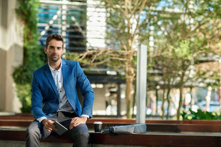 Confident businessman sitting outside his office building using a tabletの写真素材