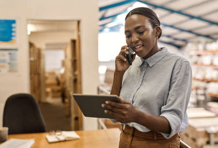 Smiling African woman using a phone and tablet in a warehouseの写真素材