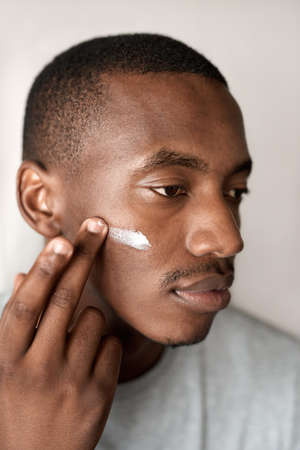 Young African man applying moisturizing cream to his cheekの写真素材