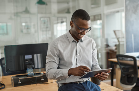 Smiling African businessman leaning on an office desk using a tabletの写真素材