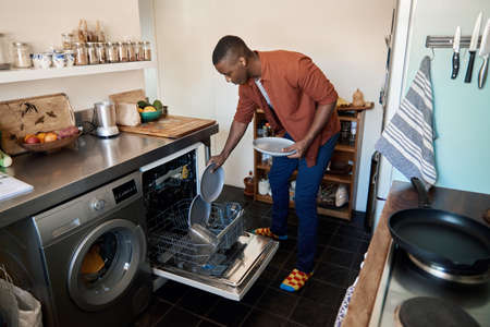 Young African man putting some plates in a dishwasher at homeの写真素材