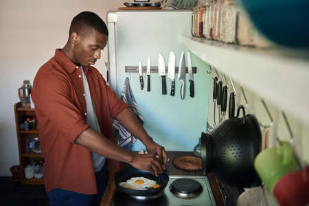 Young African man cracking eggs into a frying panの写真素材