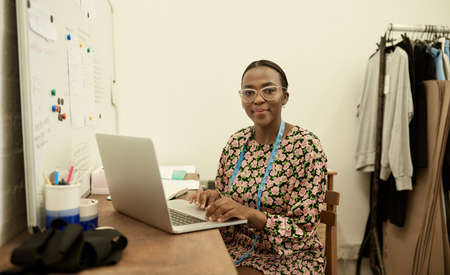 Smiling African female fashion designer working on laptop in her studioの写真素材