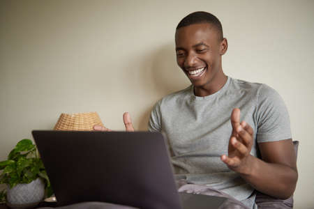 Laughing young African man using a laptop in bed at homeの写真素材