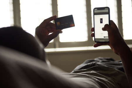 Young African man shopping online in bed in the morningの写真素材