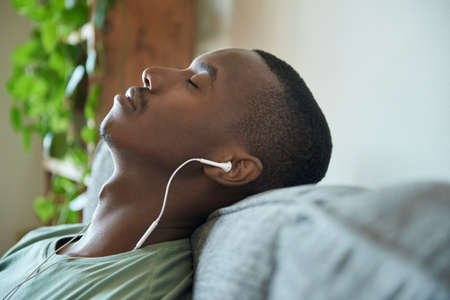 Young African man listening to music on earphones on his sofaの写真素材