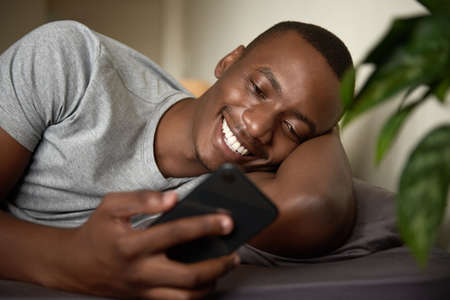 Smiling African man reading a text in bed in the morningの写真素材