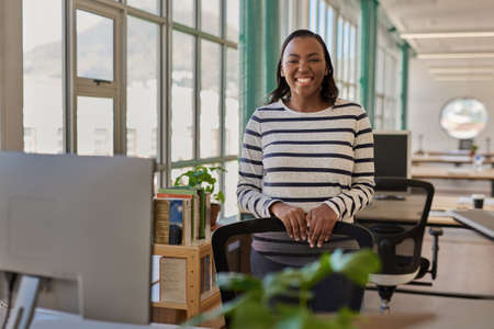 Smiling young African businesswoman standing by her desk in an officeの写真素材