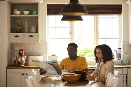 Young multiethnic couple catching on news during breakfast in their kitchenの写真素材