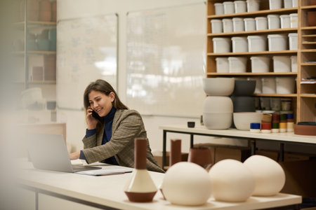 Smiling ceramics studio manager talking on a phone and using a laptopの写真素材