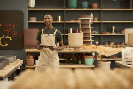 Young African potter with a tablet smiling in a large ceramics workshopの写真素材