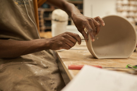 African ceramist making holes in a pot at a workbench in a studioの写真素材