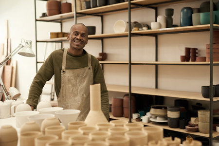 Smiling African ceramist standing by a bench and shelves full of potsの写真素材