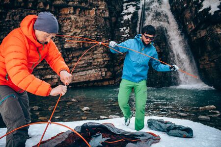 Two young professional male tourists are preparing to camp in the mountains near the river in winter. White waterfall and beautiful texture of rocks on background.
Travel, trekking  and active life concept with team. Hiking, camping equipment.の写真素材