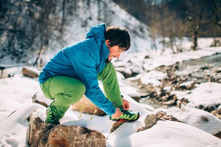 Young male tourist ties shoe laces near a river in the mountains.
Beautiful winter landscape with snow covered banks and trees on background. Climbing, trekking, active life, camping concept.の写真素材