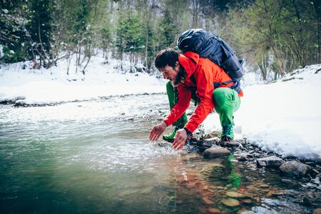 Young male tourist washes his face with river water in the mountains.
Beautiful winter landscape with snow covered banks and trees on background. Climbing, hiking, trekking, active life concept.の写真素材