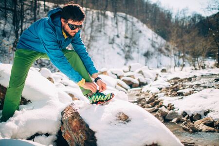 Young male tourist ties shoe laces near a river in the mountains.
Beautiful winter landscape with snow covered banks and trees on background. Climbing, trekking, active life, camping concept.の写真素材