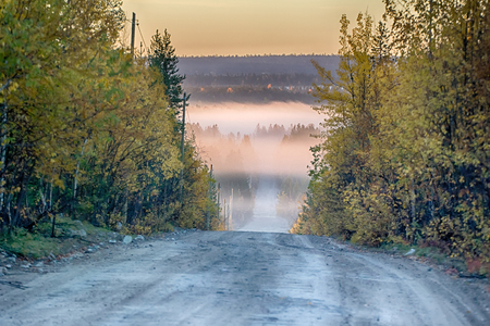 Scenic route, car trip. Dirt road through autumn woods, fog illuminated by the sunの写真素材