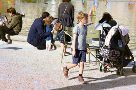 Paris, France - September 23, 2017: blond hair boy (pretty boy, girl-boy) with stick (wand to control ships in pool) in Park where people walk with children, city fountain. Street lifeのeditorial素材