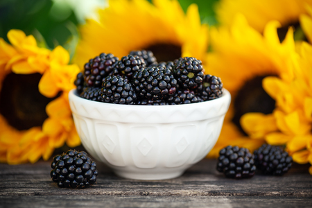 Ripe blackberries in white bowl with sunflower bouquet on wooden table, summer theme.の写真素材
