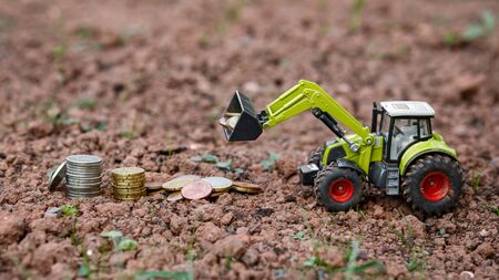 Closeup Of Small green Agricultural Toy Tractor loading money coins, Icon Farmer Machinery,...