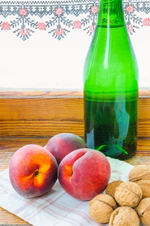Three red peaches, a green glass bottle of wine and some walnuts, close to the windowの写真素材