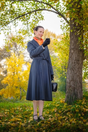 Senior business woman with a mobile phone, a bag and an orange scarf, walking in the park, under the trees in autumnの写真素材