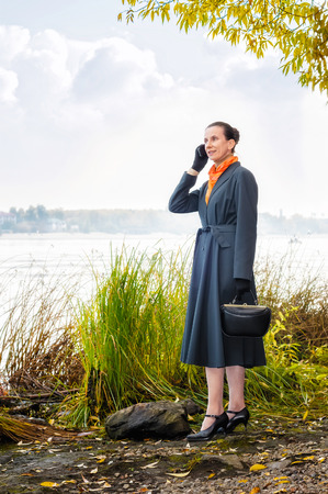 Elegant senior business woman with a mobile phone, a bag and an orange scarf, walking along the river, under the trees in autumnの写真素材
