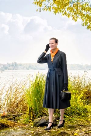 Elegant senior business woman with a mobile phone, a bag and an orange scarf, walking along the river, under the trees in autumnの写真素材