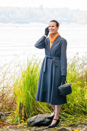 Elegant senior business woman with a mobile phone, a bag and an orange scarf, walking along the river, under the trees in autumnの写真素材