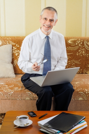 An handsome senior businessman working with computer at home, holding spectacles in his hand and ready to drink a hot cup of black coffeeの写真素材