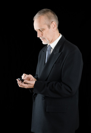Senior businessman in black suit writing sms or dialing a number on his mobile phone, on black backgroundの写真素材