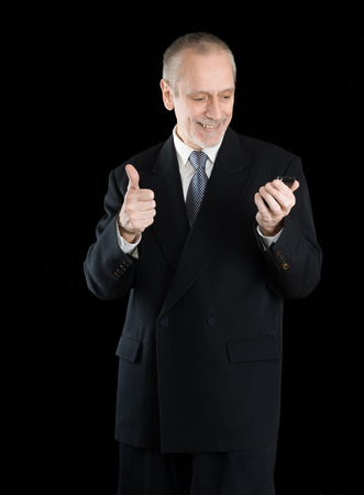 An amiable businessman wearing a black suit  smiling and reading on mobile phone, with thumb up, on black backgroundの写真素材