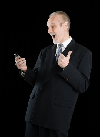 Happy businessman wearing a black suit, smiling and reading sms on mobile phone, with thumb up, on black backgroundの写真素材