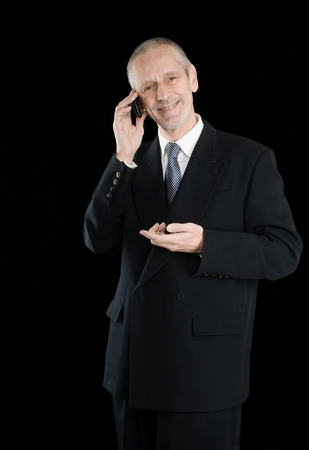 An agreeable businessman wearing a black suit smiling while speaking on mobile phone, on black backgroundの写真素材