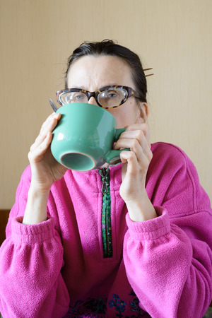 A mature woman with glasses wearing a pink pull-over and drinking coffee in a green cup at morning for breakfastの写真素材
