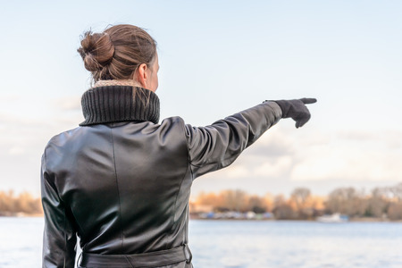 An adult with a chignon and wearing a black leather coat stays close to the river and point her finger to indicate something interesting on the opposite bankの写真素材