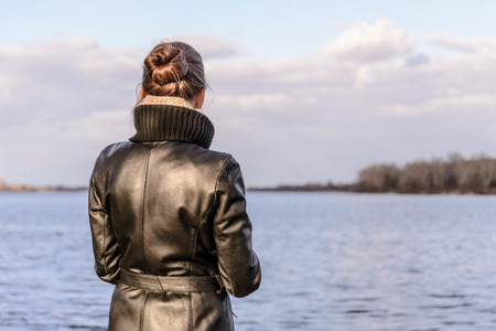 A woman with a chignon and a black leather coat is watching the landscape close to the lake or the river during a sunny day with big white cloudsの写真素材