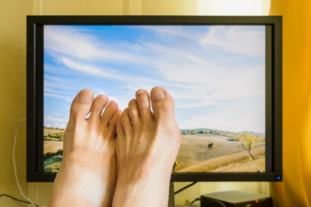 Man's feet in front of a computer monitor with a nice sunny italian landscape with a cloudy sky to symbolize the wish of holidays restの写真素材