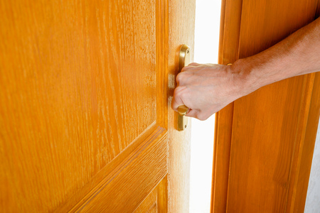 A man is opening a wooden door with a metallic handle to get out of a room and enter in an illuminated one.の写真素材