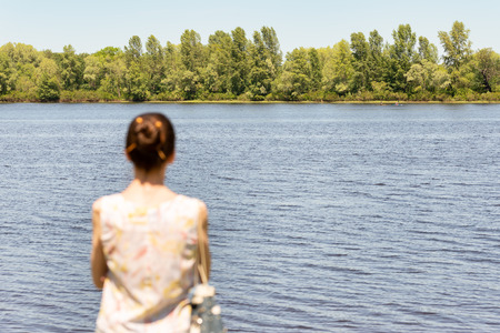 A woman with a chignon, standing up close to the Dnieper river in Kiev, Ukraine, observes the trees in the distance. The silhouette of the lady is out of focus, against a focused background.の写真素材