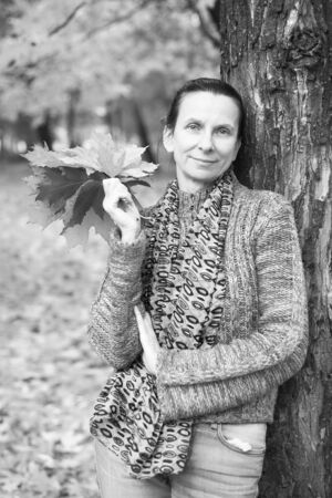 Black and white portrait of a smiling adult caucasian woman is picking yellow maple leaves in autumn in the parkの写真素材