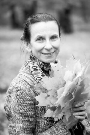 Black and white portrait of a smiling adult caucasian woman is picking yellow maple leaves in autumn in the parkの写真素材