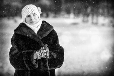 A winter portrait of a smiling senior adult woman wearing a wool cap, a scarf and colored gloves, with a snow backgroundの写真素材