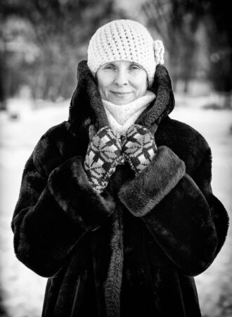 A winter portrait of a smiling senior adult woman wearing a wool cap, a scarf and colored gloves, with a snow backgroundの写真素材